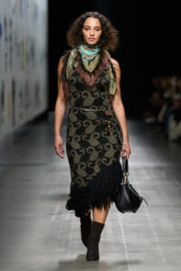 Fashion model walking a runway in a sleeveless patterned dress with fringe hem, green scarf, and a chain belt, carrying a black handbag.