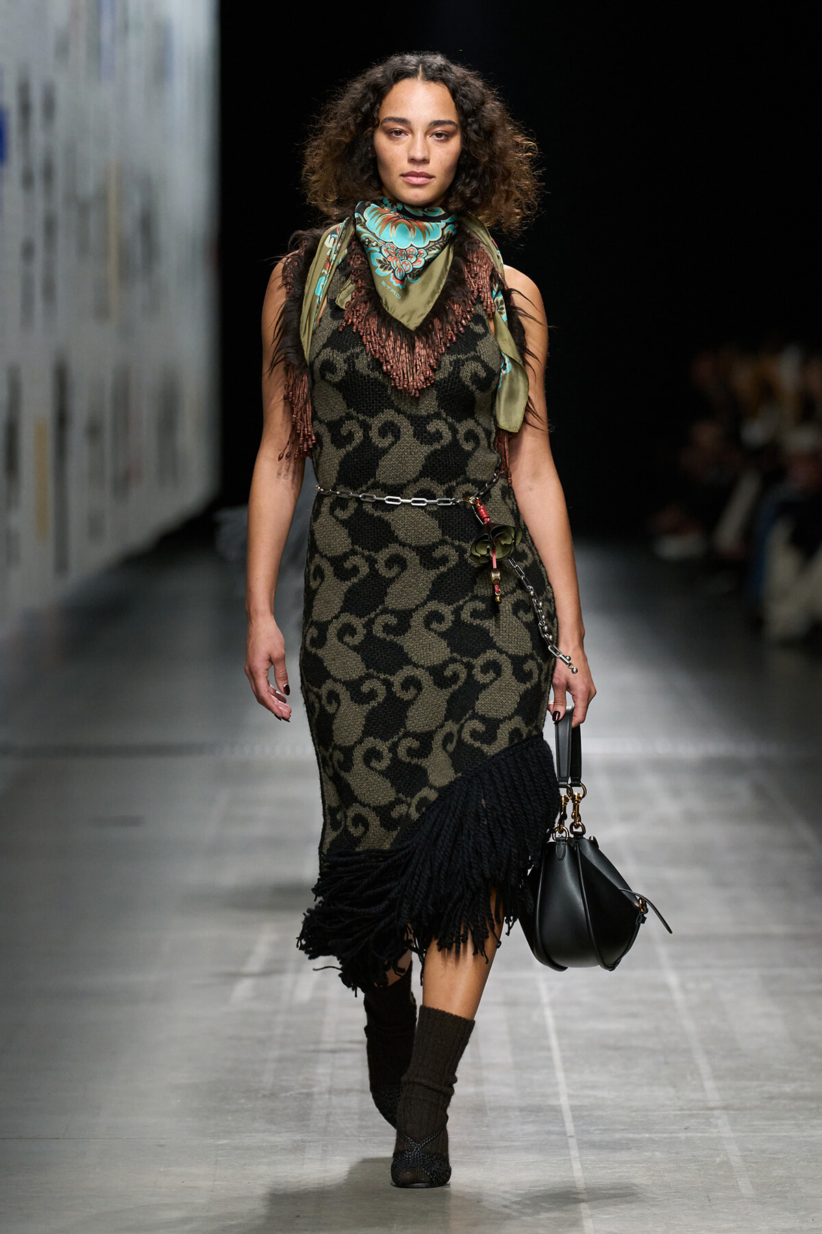 Fashion model walking a runway in a sleeveless patterned dress with fringe hem, green scarf, and a chain belt, carrying a black handbag.