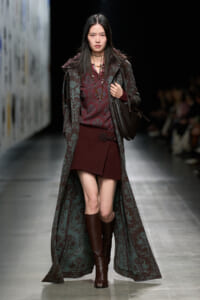 Model on a runway wearing a burgundy outfit: knit top and short skirt, long teal floral coat, brown knee-high boots, and a brown shoulder bag