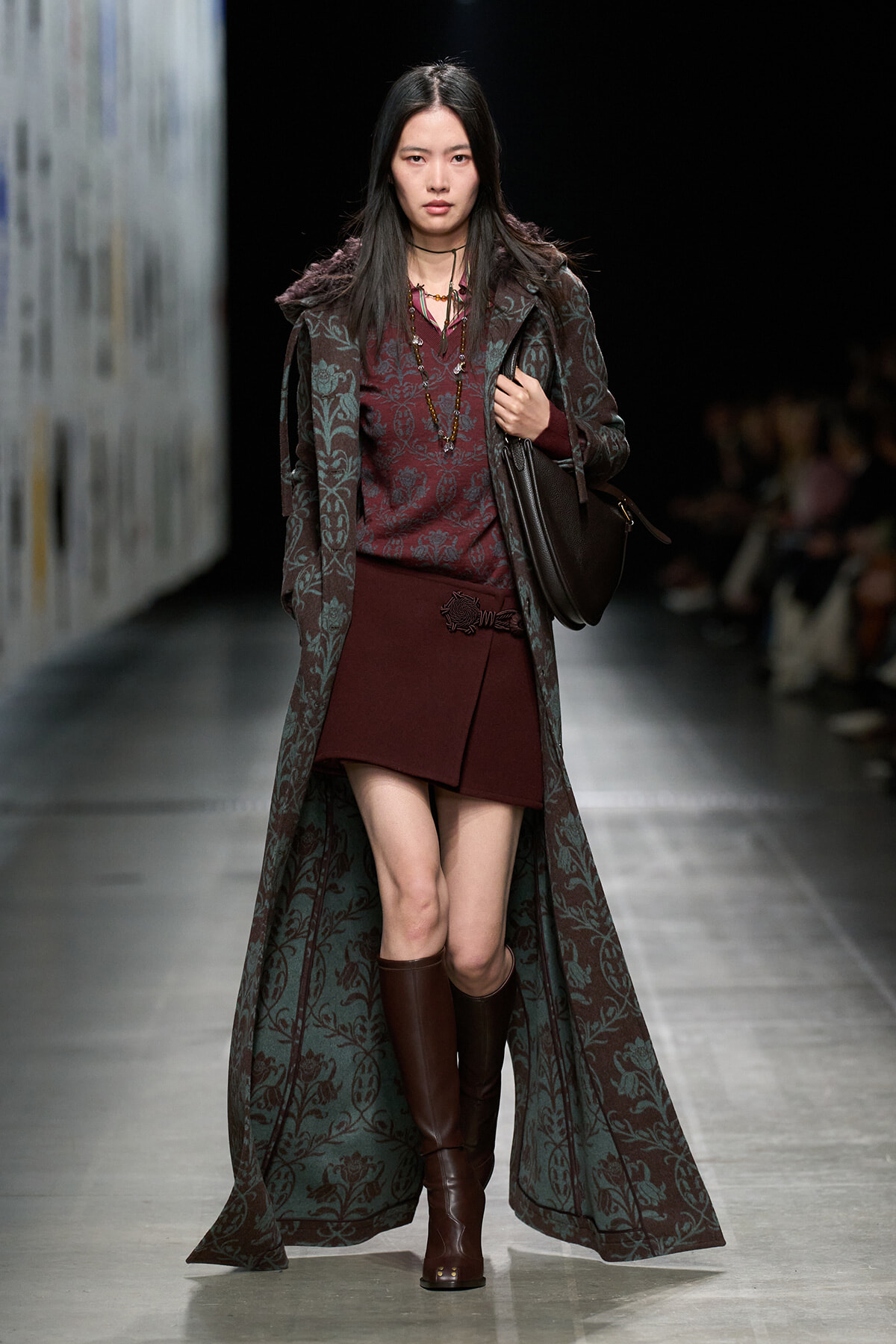 Model on a runway wearing a burgundy outfit: knit top and short skirt, long teal floral coat, brown knee-high boots, and a brown shoulder bag