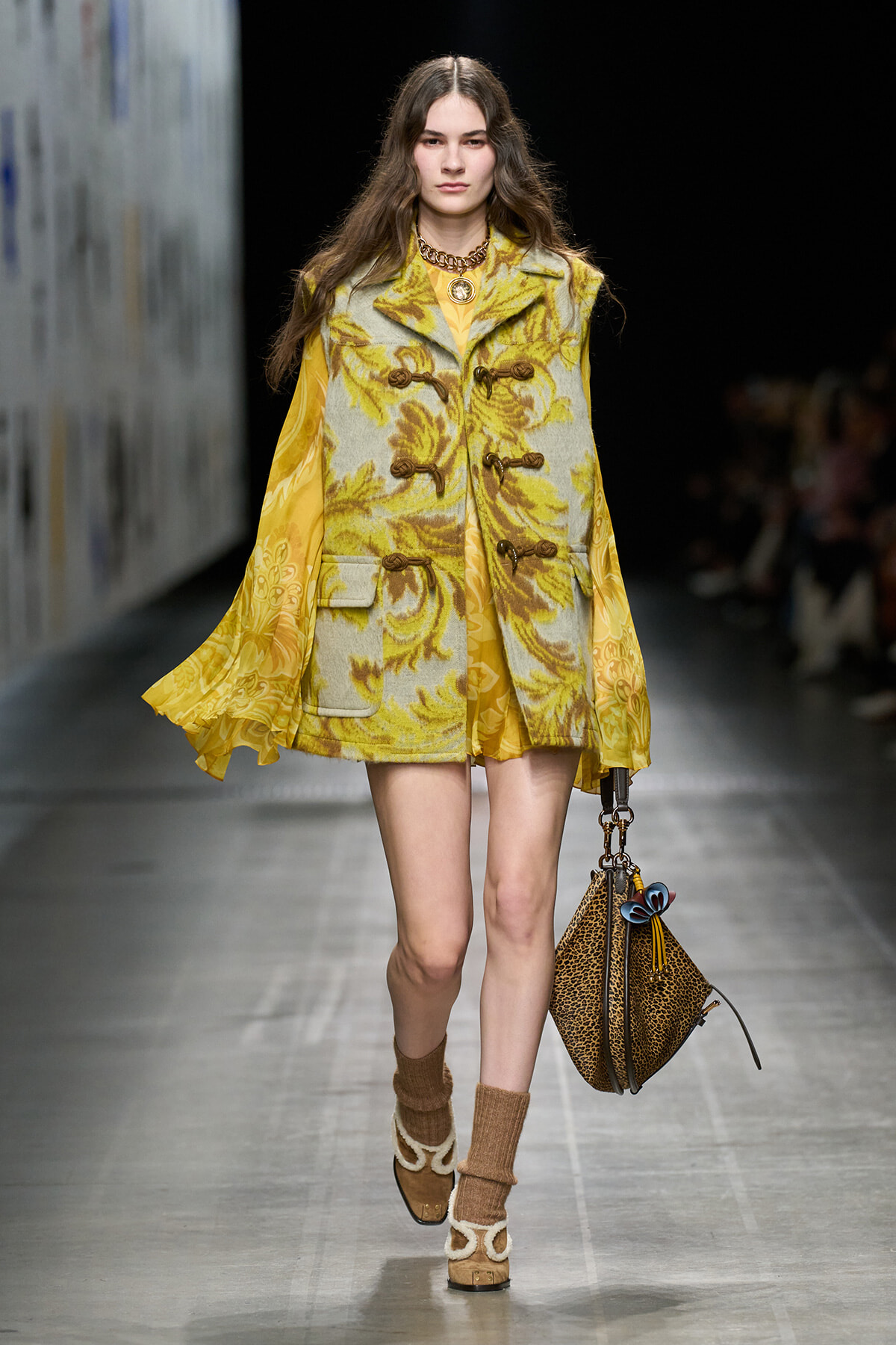 Model walking the runway in a yellow floral sleeveless coat with toggle closures over a yellow dress, paired with leopard print bag and chunky boots.