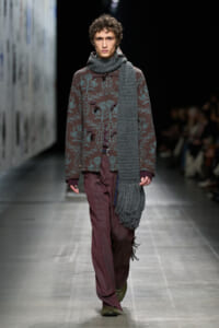 Male model walking a runway in a dark teal floral-patterned coat with a large gray scarf, burgundy pinstriped pants, and green shoes, spotlight on him