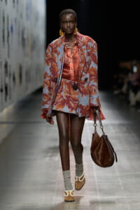 Model on a runway wearing a lavender floral coat over a coral satin dress, with large gold earrings and a brown leopard-print handbag.