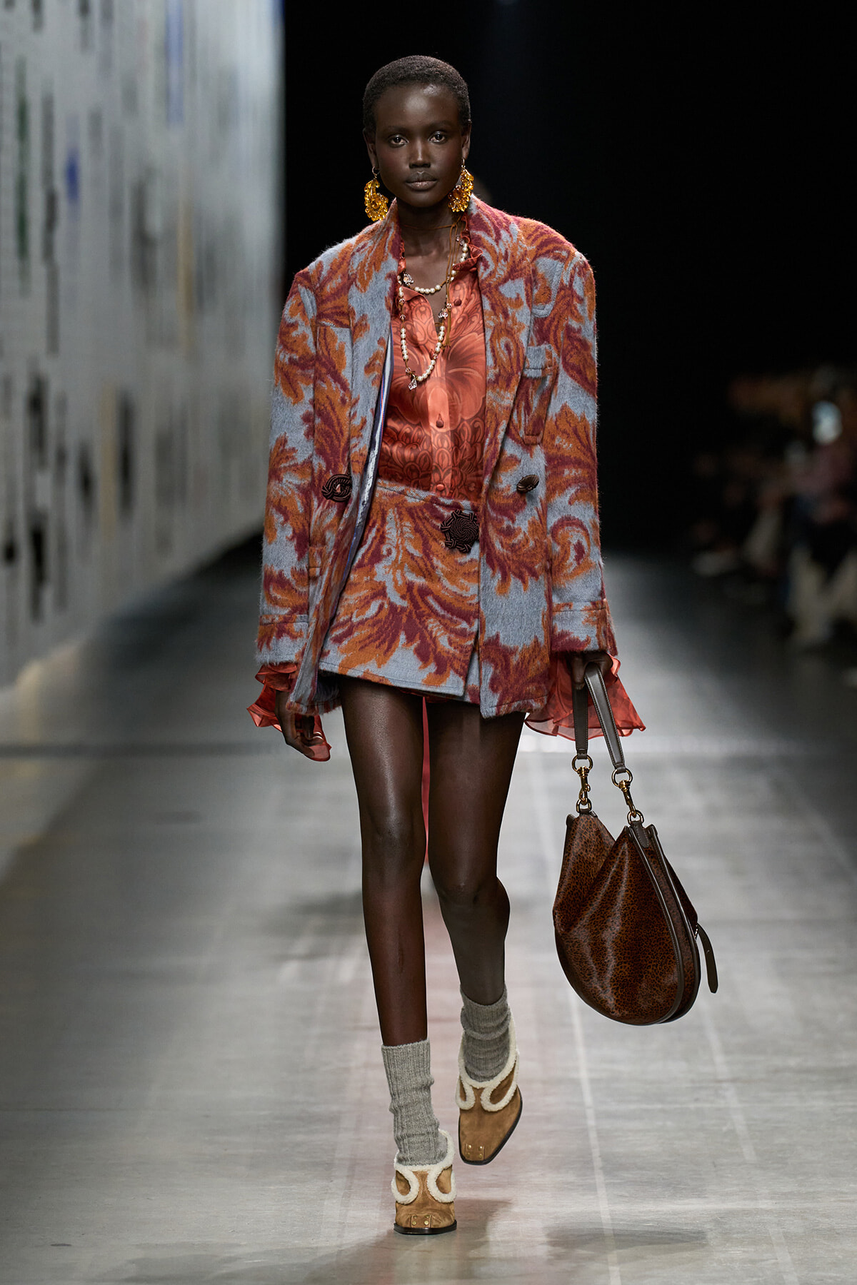 Model on a runway wearing a lavender floral coat over a coral satin dress, with large gold earrings and a brown leopard-print handbag.