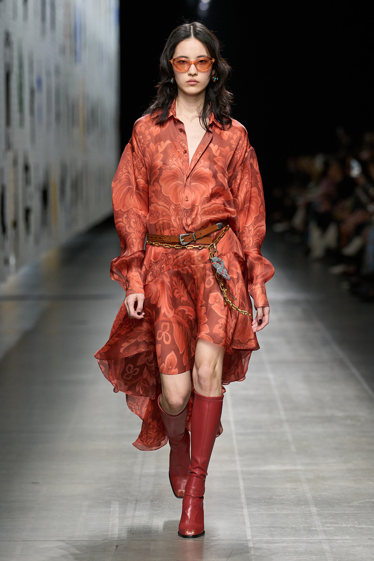 Model walking runway in a red floral-pattern shirt dress with a high-low hem, brown belt, and chunky red boots with orange sunglasses