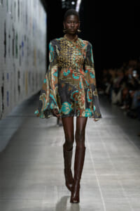 Runway model walking in a colorful floral-print dress with a dramatic gold chain chest harness and tall brown boots.