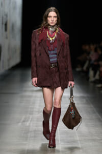 Model on a runway wearing a burgundy patterned coat, matching scarf, knee-high burgundy boots, and a brown handbag.