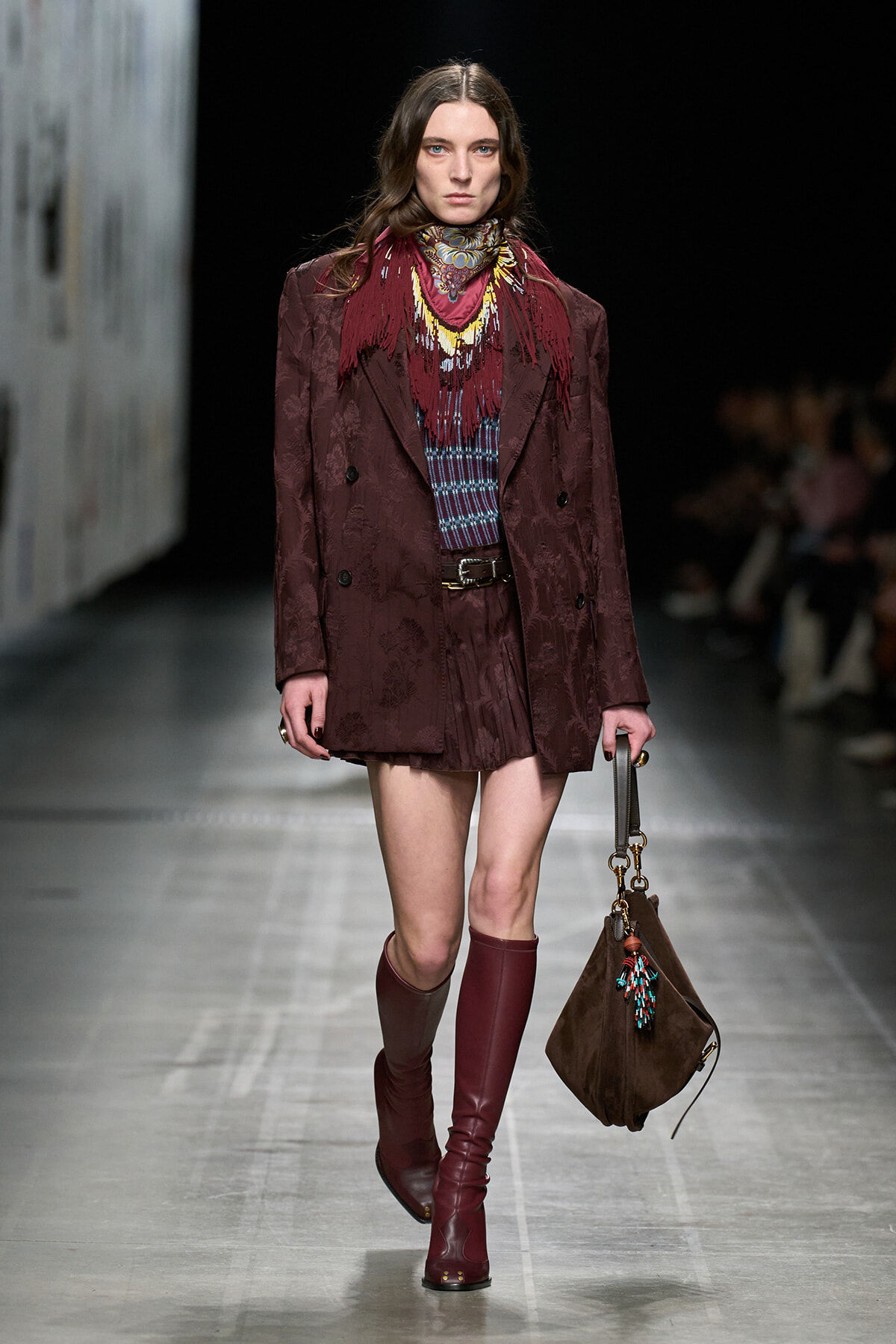 Model on a runway wearing a burgundy patterned coat, matching scarf, knee-high burgundy boots, and a brown handbag.