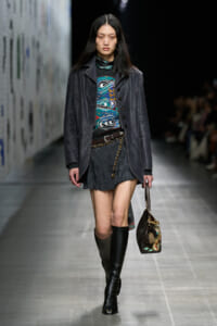 Female model walks the runway in a dark leather coat over a colorful patterned sweater, black mini skirt, tall black boots, and a chain belt, carrying a patterned handbag.