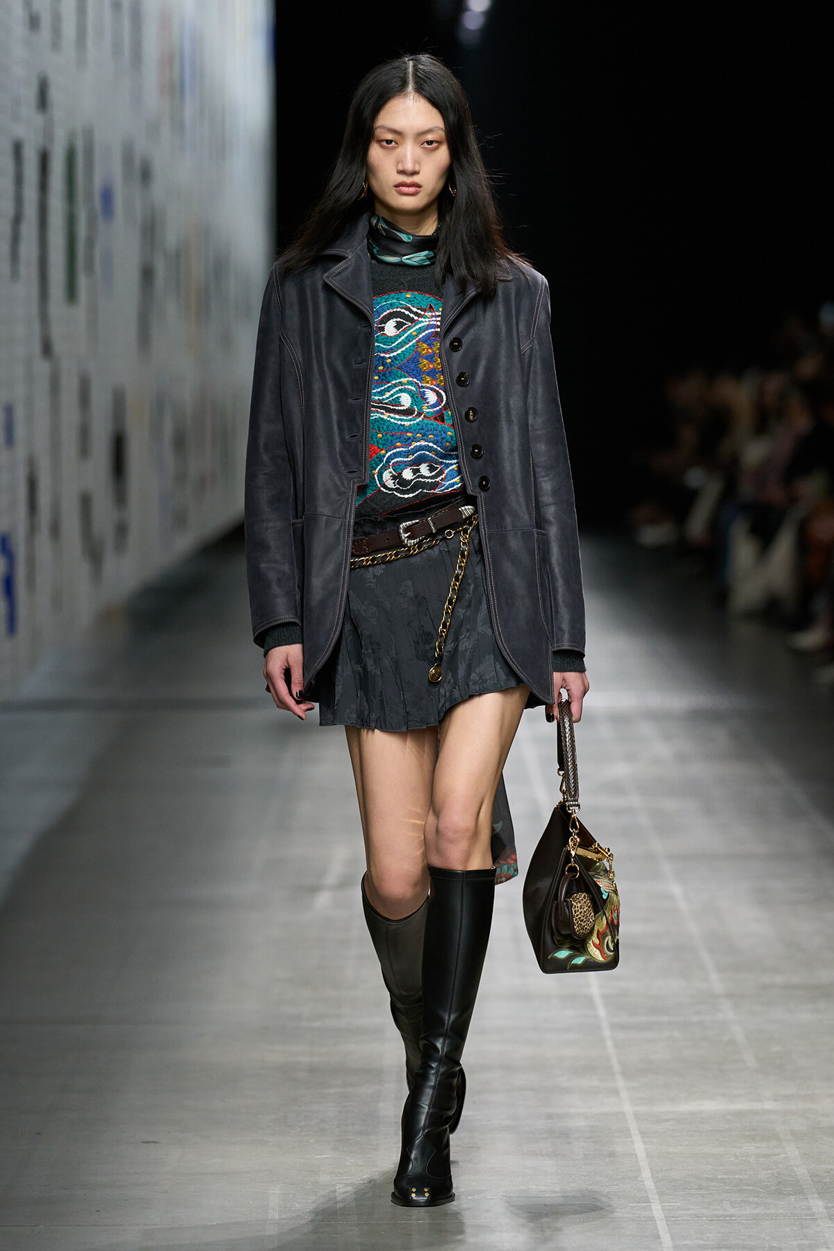 Female model walks the runway in a dark leather coat over a colorful patterned sweater, black mini skirt, tall black boots, and a chain belt, carrying a patterned handbag.