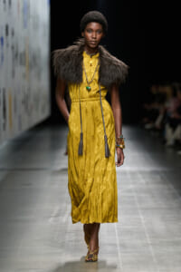 Runway model in a mustard-yellow pleated midi dress with a shaggy fur shoulder cape, layered necklaces, and braided sandals.