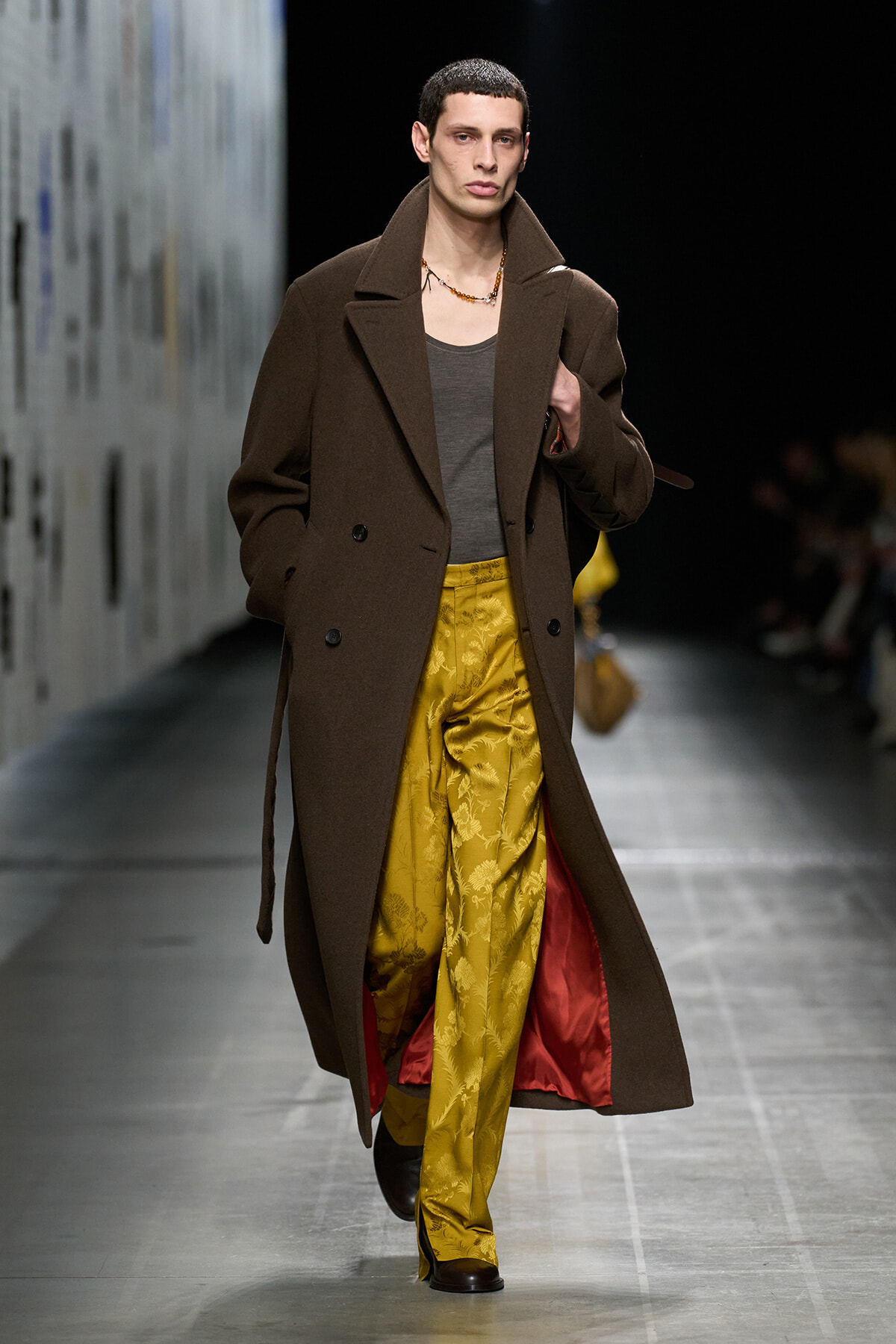 Male model on runway wearing a long brown coat, gray tee, and gold brocade pants with a beaded necklace.
