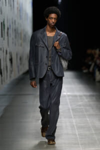 Male model walking a runway in a dark blue-gray suit with a matching blazer, gray shirt, and a shoulder bag strapped on his back.