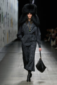 Model walking a runway in a dark, patterned long dress with a large black fur hat and gold necklace, carrying a black handbag.