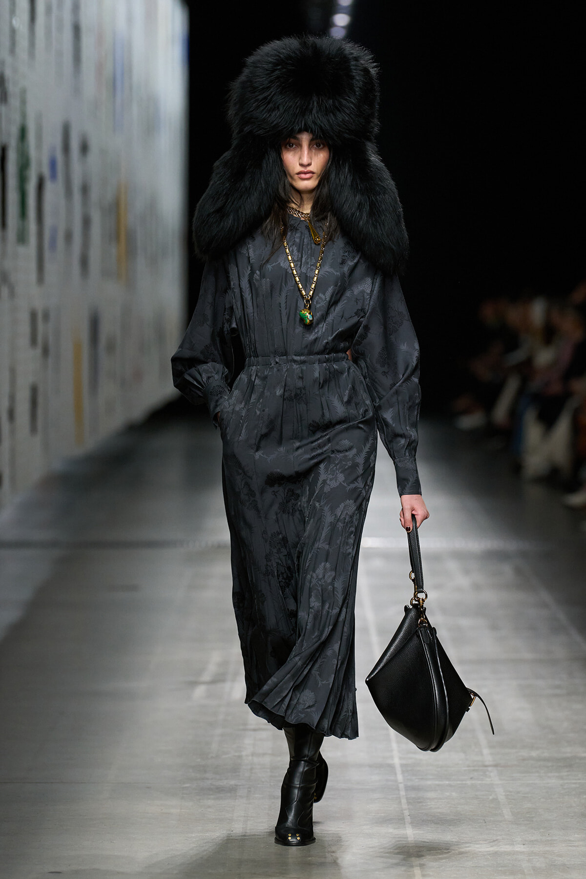 Model walking a runway in a dark, patterned long dress with a large black fur hat and gold necklace, carrying a black handbag.