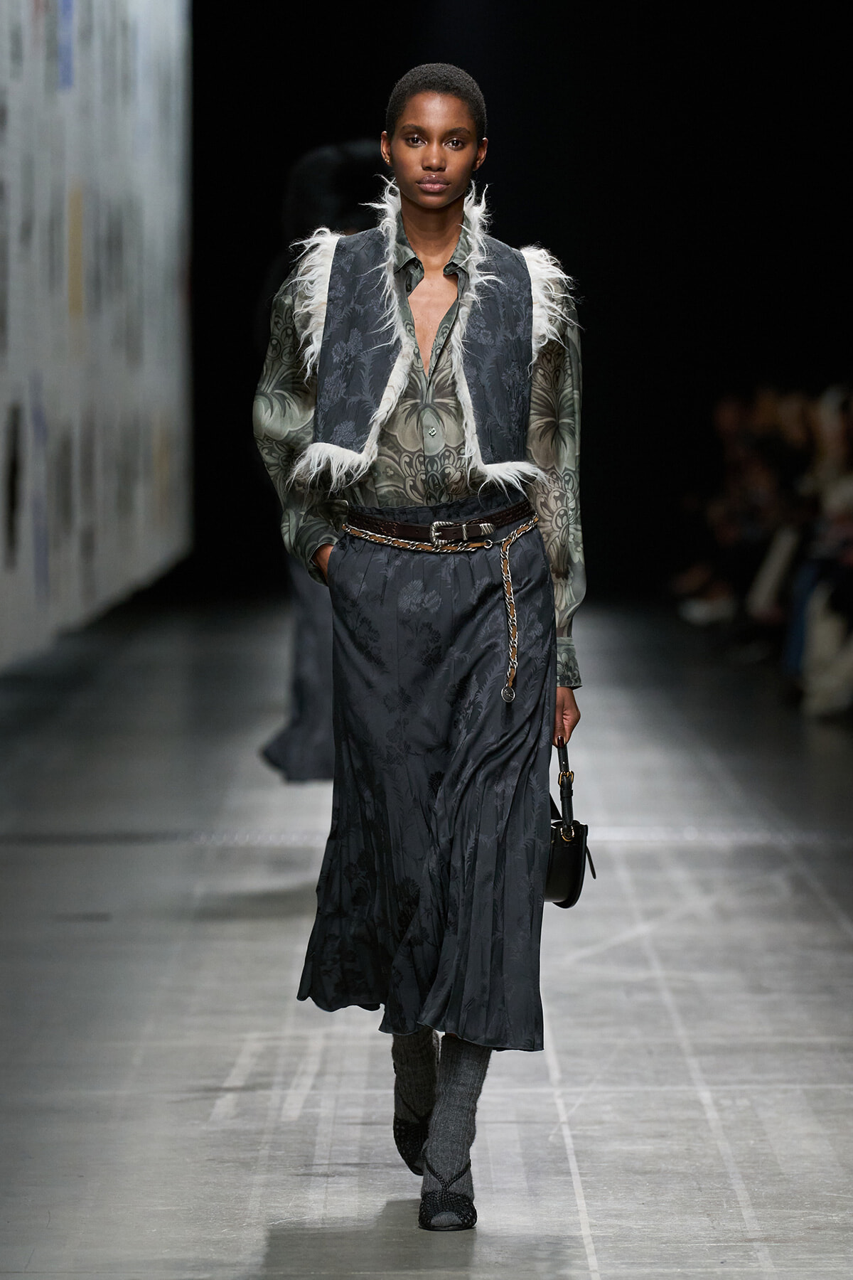 Model walks runway in a dark, paisley-like blouse with a matching fur-trimmed sleeveless vest and a long skirt, accessorized with a chain belt and small black handbag