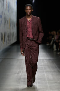 Male model walking a burgundy paisley suit on the runway with hands in pockets at a fashion show.