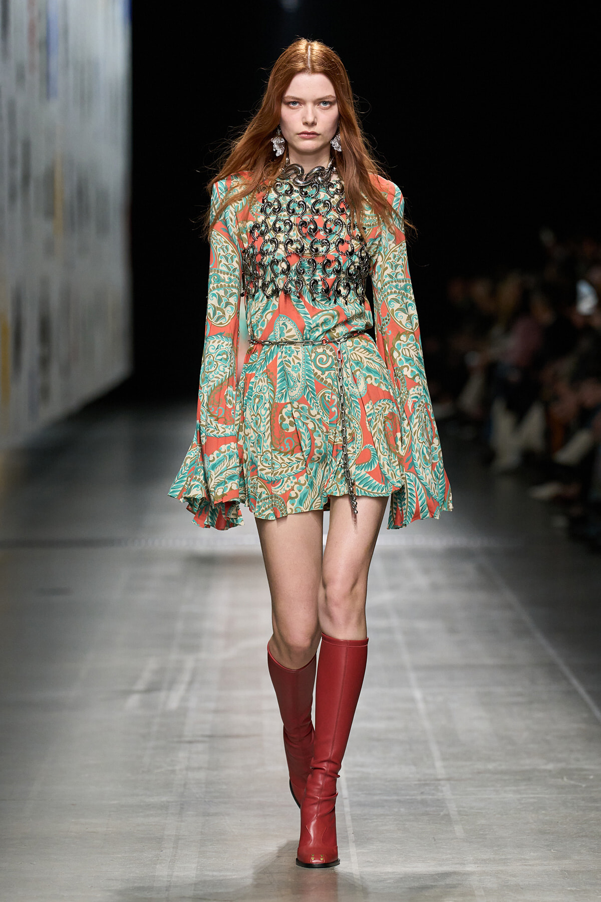 Model walking a fashion runway in a teal and coral patterned mini dress with a black lace bib and red knee-high boots.