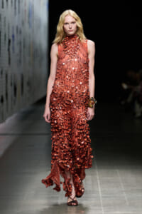 Model walks the runway in a rust-red, floor-length sequin dress with a high neckline, densely covered in circular discs, paired with bangles on her right wrist.