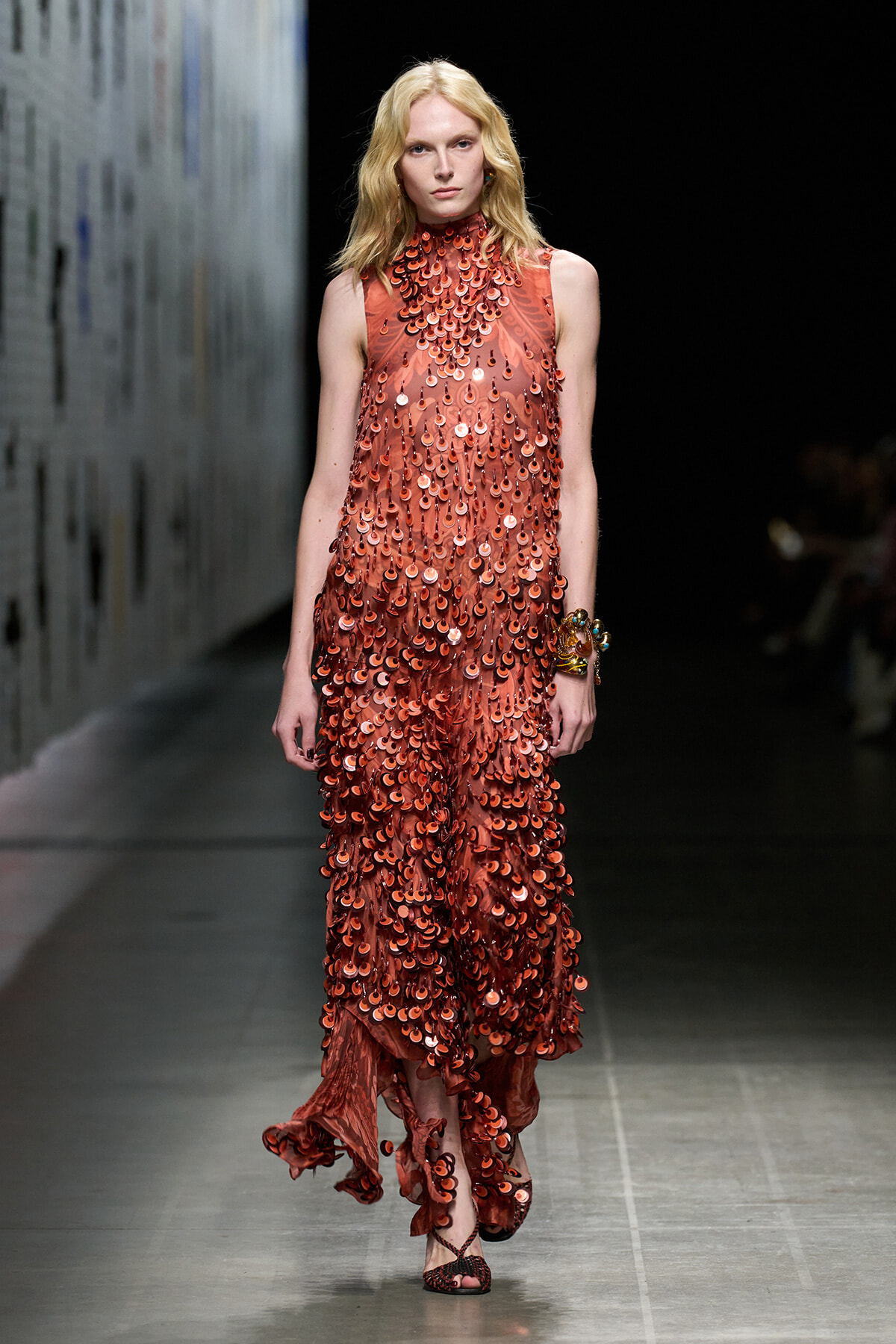 Model walks the runway in a rust-red, floor-length sequin dress with a high neckline, densely covered in circular discs, paired with bangles on her right wrist.