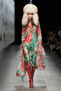 Model walks the runway in a flowing coral and teal floral dress with a large cream fur hat and red knee-high boots, accessorized with a pendant necklace.