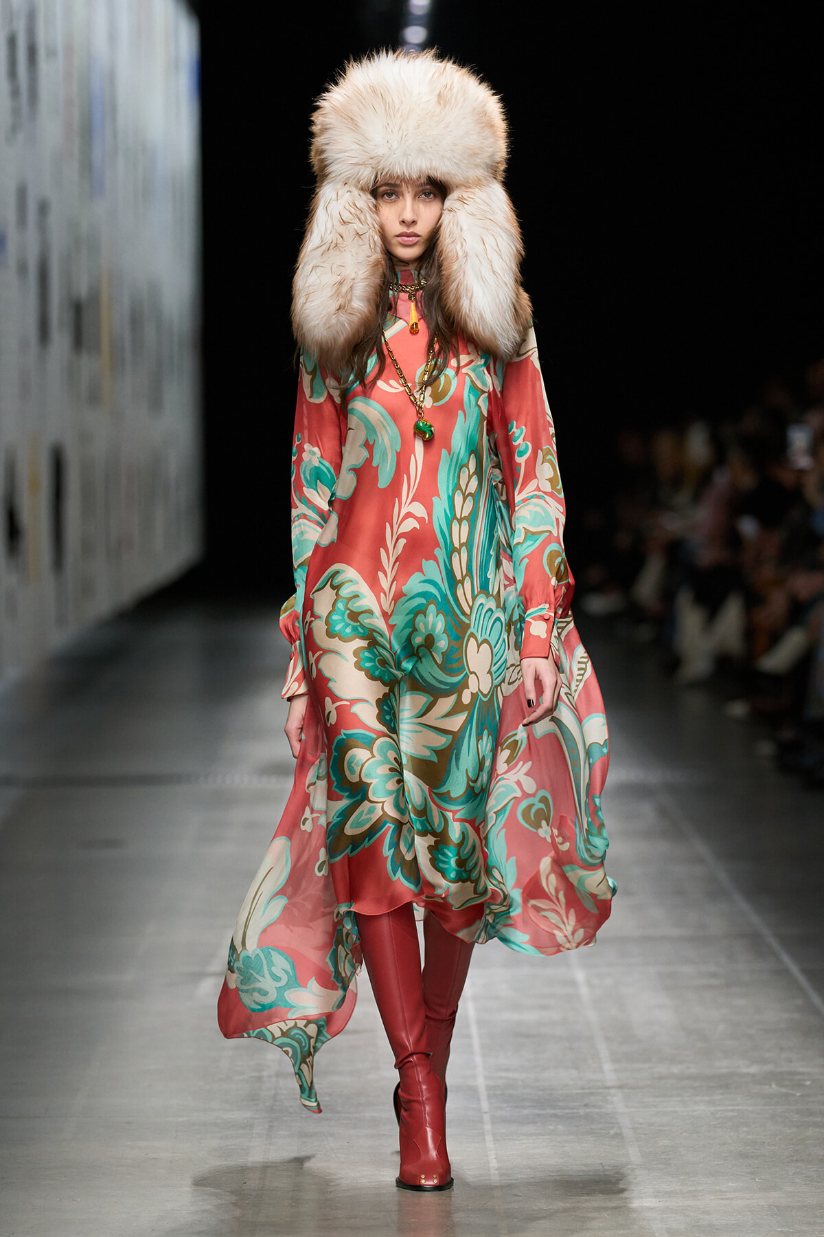 Model walks the runway in a flowing coral and teal floral dress with a large cream fur hat and red knee-high boots, accessorized with a pendant necklace.