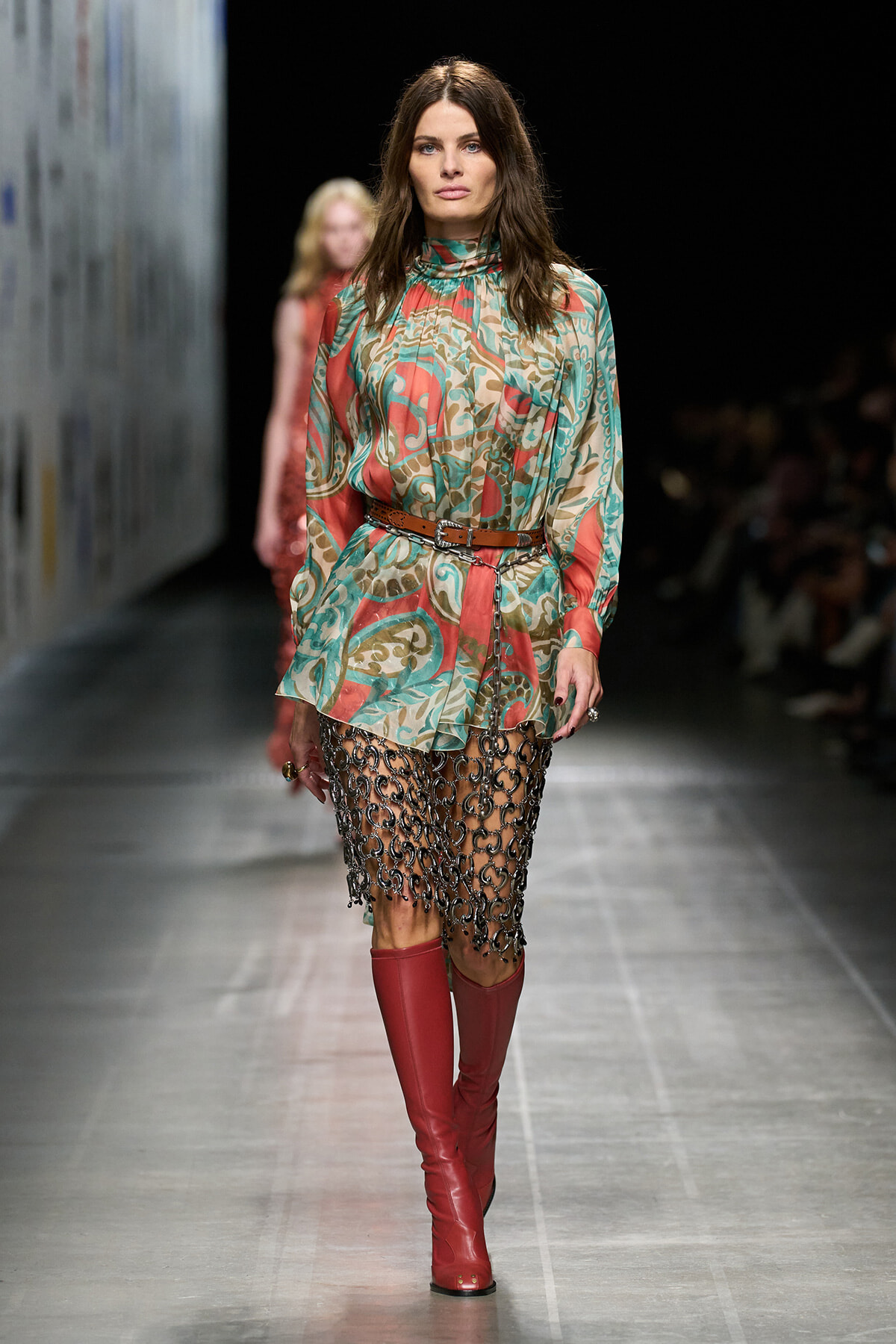 Model walks a fashion runway in a colorful, patterned tunic dress cinched with a brown belt, and a chain-link fringe skirt over red knee-high boots