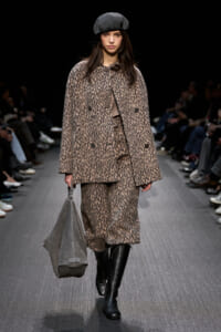 Model walking the runway in a leopard-print coat and matching pants, paired with a gray beret and knee-high black boots, carrying a large gray bag.