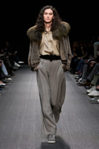 Model walks the runway in a taupe shirt tucked into high-waisted gray pleated trousers with a wide black belt and a brown fur-shouldered jacket; gray sneakers complete the look.