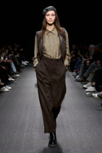 Model walks a fashion runway in a brown leather vest over a patterned beige blouse, with wide brown trousers, black boots, and a beret.