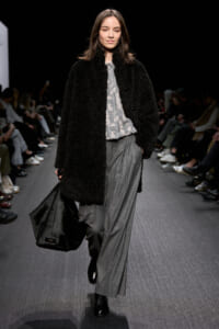 Model walks a runway in a black fuzzy coat over a gray printed blouse and wide gray pants, carrying a large black bag at her side.