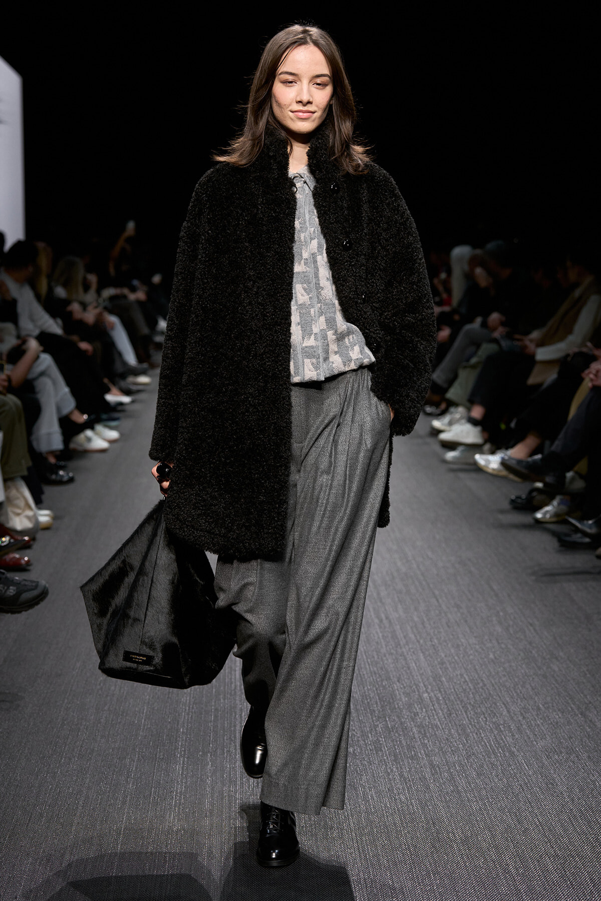 Model walks a runway in a black fuzzy coat over a gray printed blouse and wide gray pants, carrying a large black bag at her side.