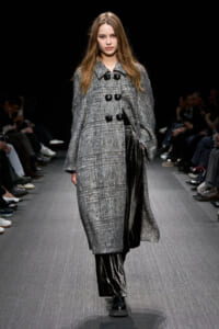 Model on a runway wearing a knee-length gray textured coat with large black toggles and black pants and boots.