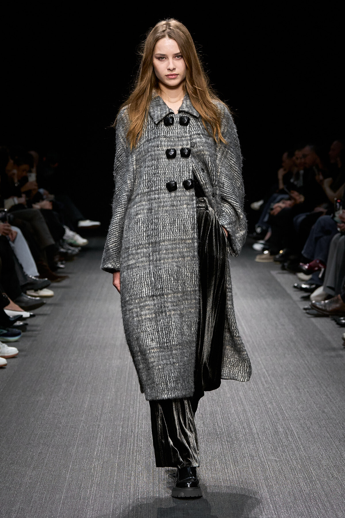 Model on a runway wearing a knee-length gray textured coat with large black toggles and black pants and boots.
