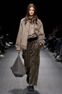 Model walks a runway in a leopard-print oversized jacket, taupe satin blouse, and high-waisted pleated metallic pants, carrying a gray slouchy bag.