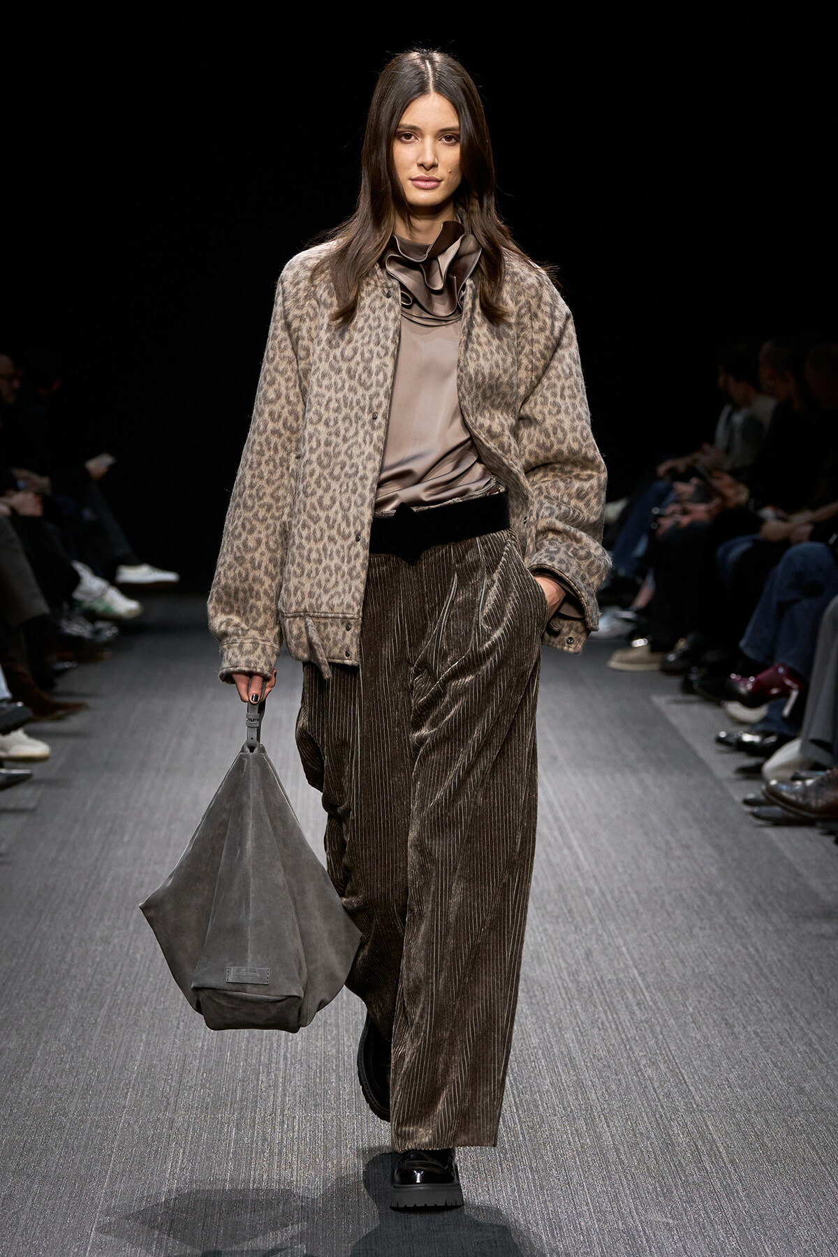 Model walks a runway in a leopard-print oversized jacket, taupe satin blouse, and high-waisted pleated metallic pants, carrying a gray slouchy bag.