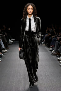 Model walks the runway in a metallic velvet pinstripe suit with a white shirt and black tie, carrying a small black clutch