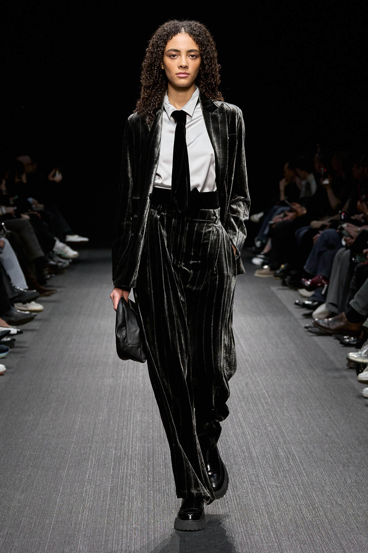 Model walks the runway in a metallic velvet pinstripe suit with a white shirt and black tie, carrying a small black clutch