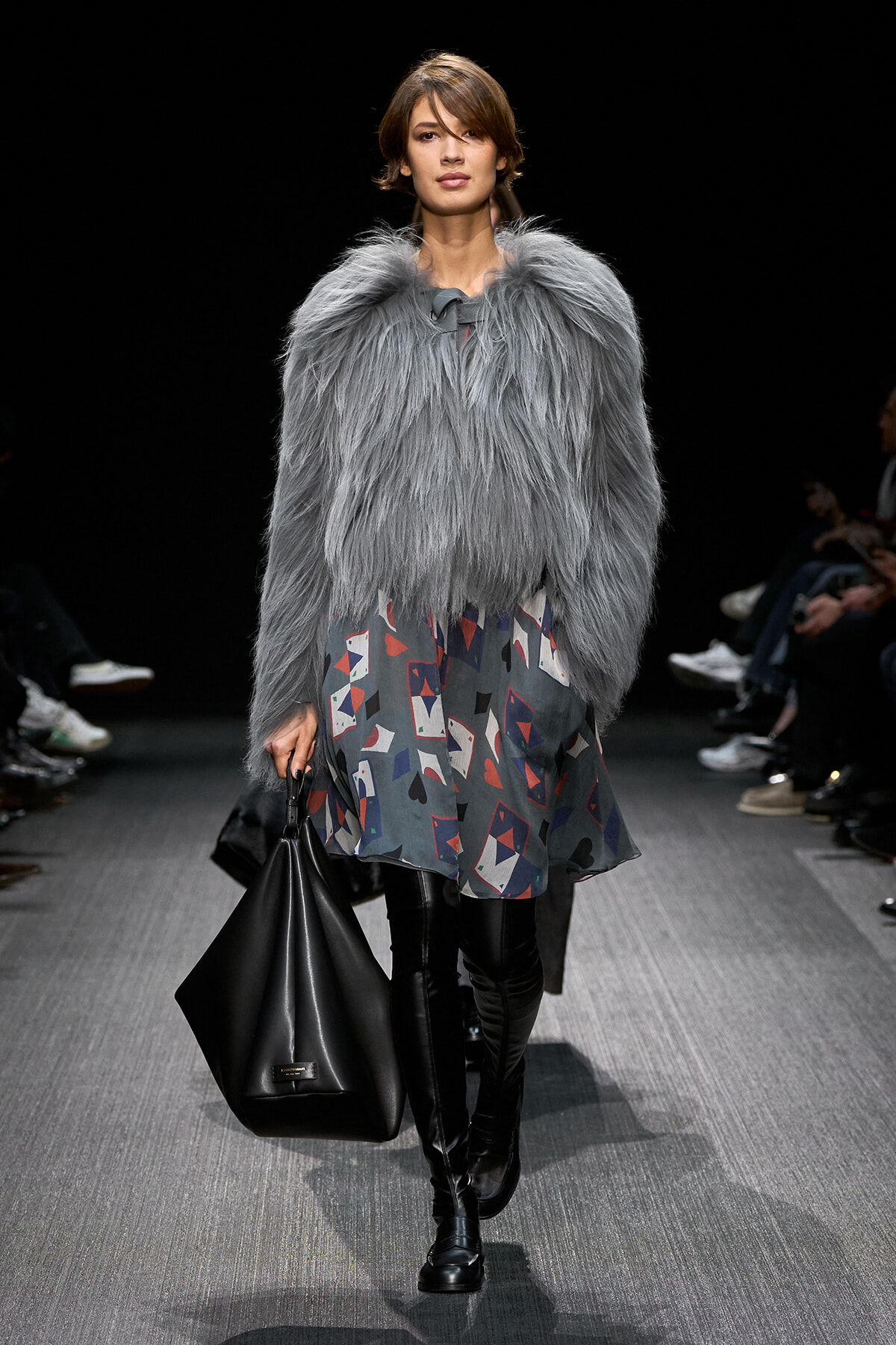 Model on a runway wearing a light gray faux-fur jacket over a geometric-print dress, paired with knee-high black boots and a large black handbag.