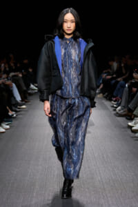 Model walking a fashion show runway in a blue marbled jumpsuit under a black textured bomber jacket with blue lining, black boots, and an attentive audience in the background.
