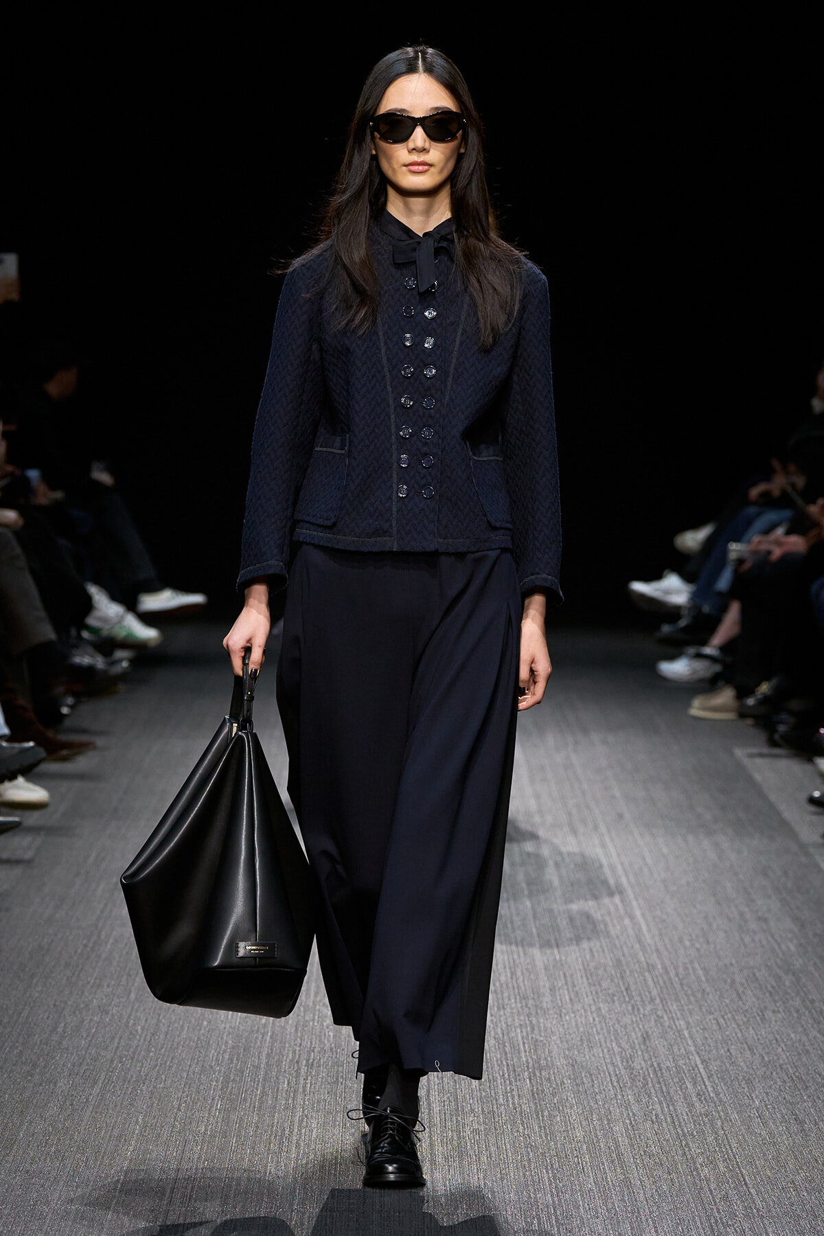 Model walks a fashion show wearing a navy textured jacket with double-button front, wide-leg navy pants, and black boots, carrying a large black tote and wearing sunglasses.