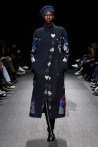 Model walks the runway in a long navy coat with playing-card suit symbols and a blue beret, black turtleneck underneath, and knee-high boots.
