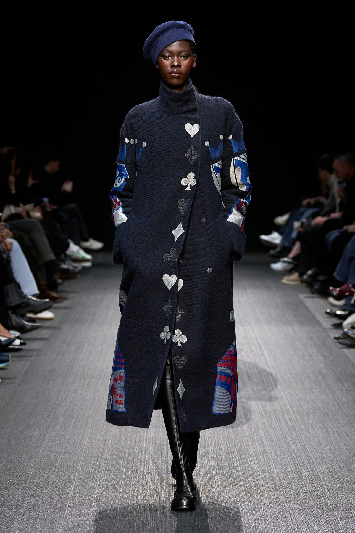 Model walks the runway in a long navy coat with playing-card suit symbols and a blue beret, black turtleneck underneath, and knee-high boots.