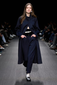 Model walking a fashion show in a navy coat and matching wide-leg trousers with a white belt, black patent shoes, and white socks.