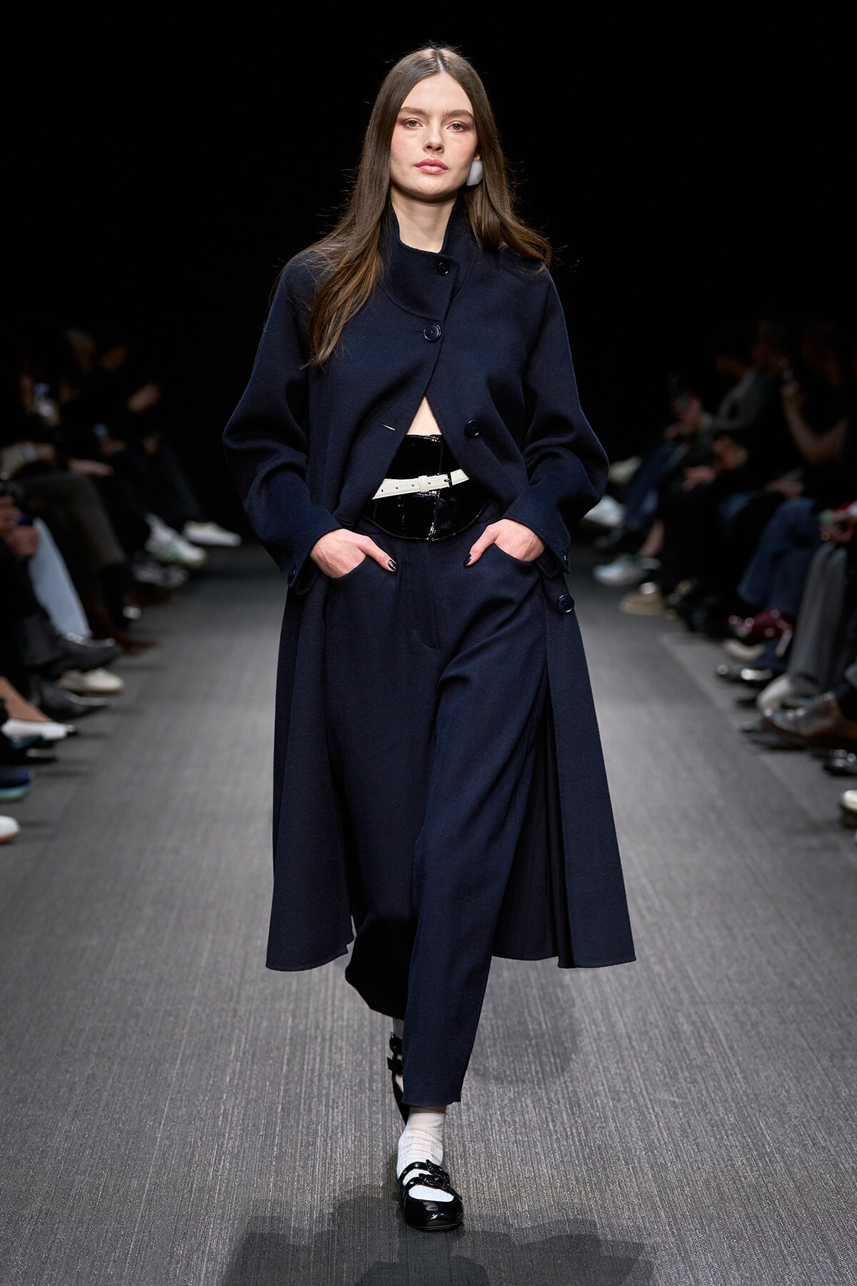 Model walking a fashion show in a navy coat and matching wide-leg trousers with a white belt, black patent shoes, and white socks.