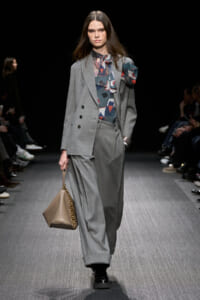 Model walks the runway in a light gray oversized suit with a patterned blouse and bow scarf, carrying a beige handbag with a gold chain strap.