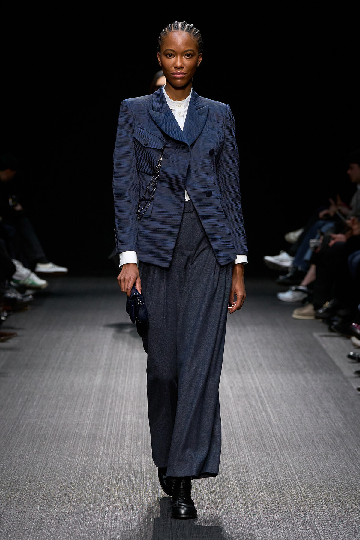 Model walking a fashion runway in a navy textured blazer over a white shirt, with wide-leg dark trousers and a small navy handbag.