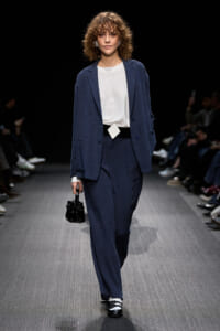 Model walks runway in a navy suit with oversized blazer and matching trousers, white blouse, square belt buckle, and black handbag.