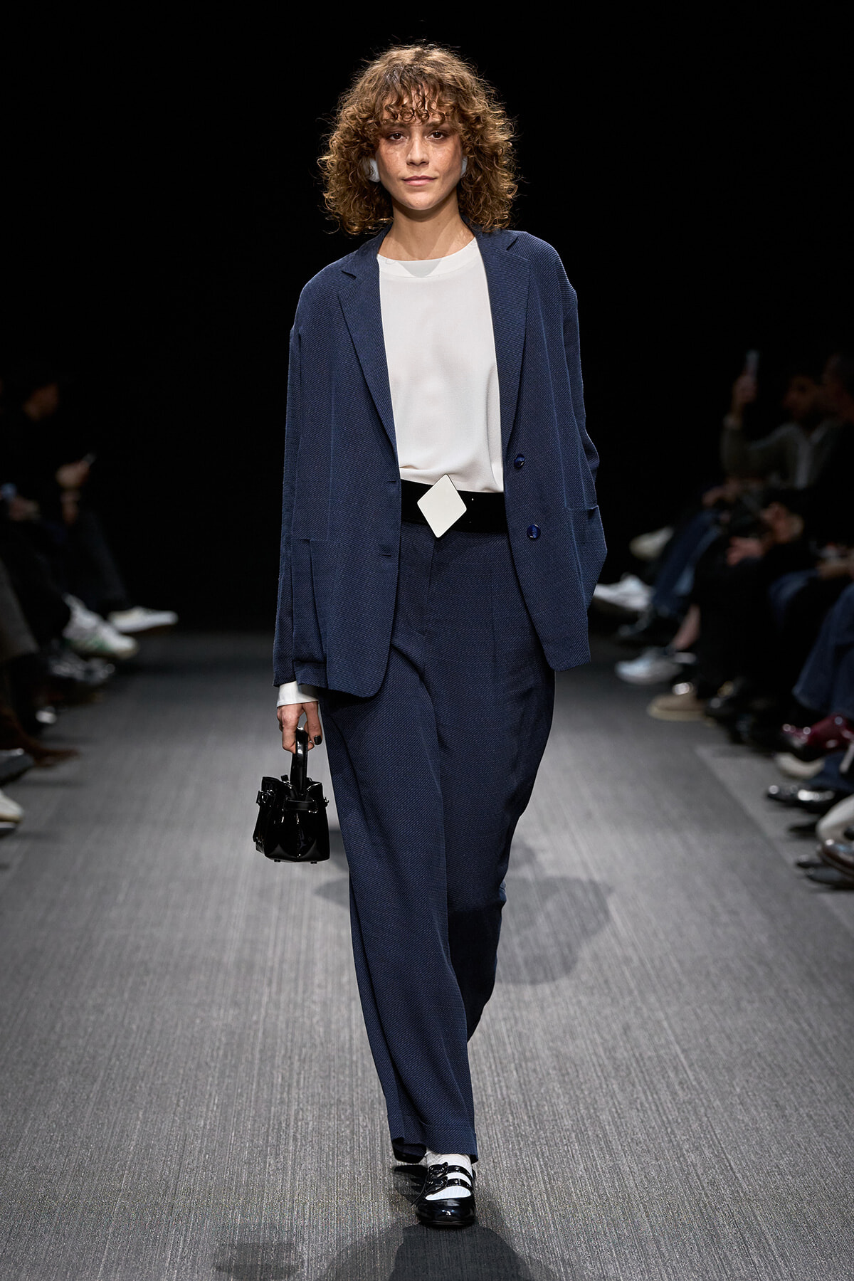 Model walks runway in a navy suit with oversized blazer and matching trousers, white blouse, square belt buckle, and black handbag.