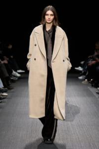 Model on a runway wearing a long beige teddy coat over a dark outfit, walking toward the camera.